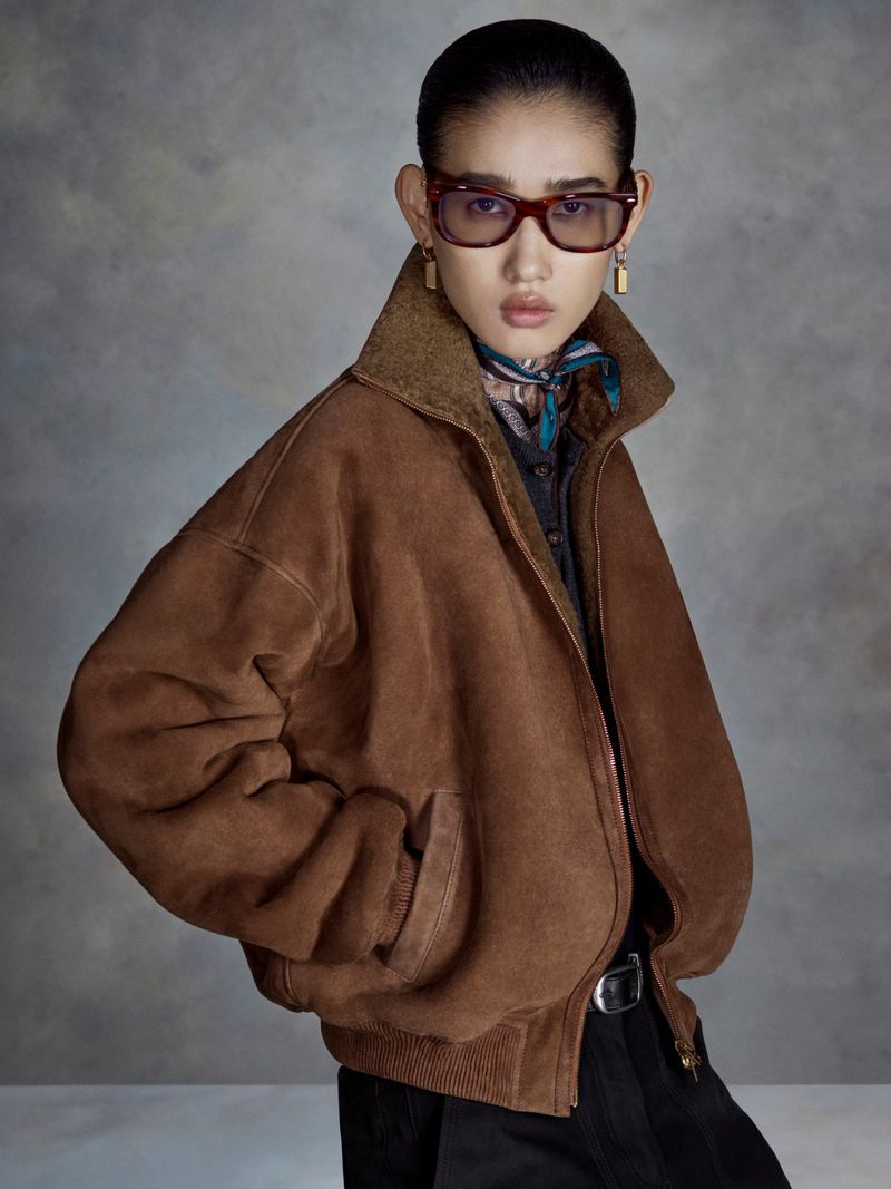 Model wearing an oversized brown suede jacket, tinted eyeglasses, and a neck scarf against a textured grey backdrop.