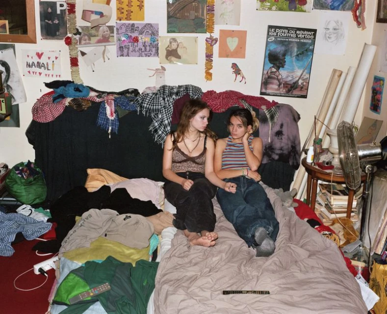 Two young women are casually posed on a bed amid cluttered bedroom, featuring posters and drawings on the wall, with natural lighting.