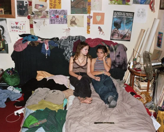 Two young women are casually posed on a bed amid cluttered bedroom, featuring posters and drawings on the wall, with natural lighting.