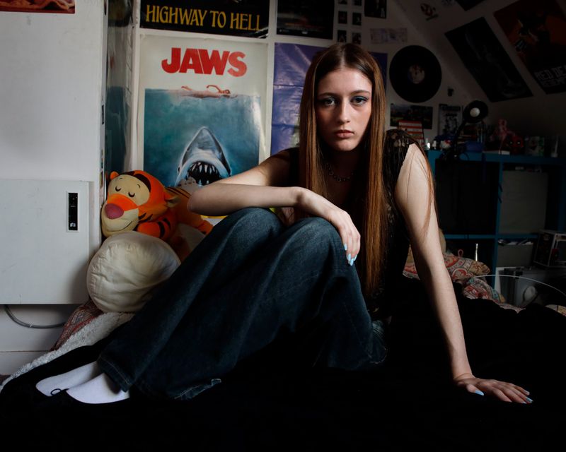 Young woman in jeans and black top sits on a black surface in a room with posters and toys, creating a casual portrait indoors.