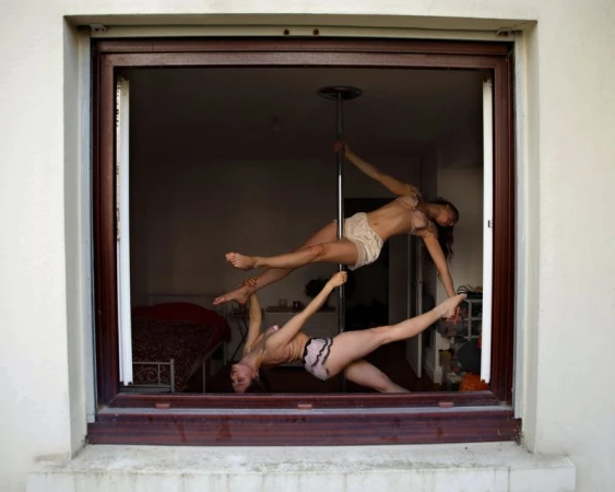 Two women in lingerie perform a pole dance routine inside a room, seen through a window. The women strike poses on and around the pole.