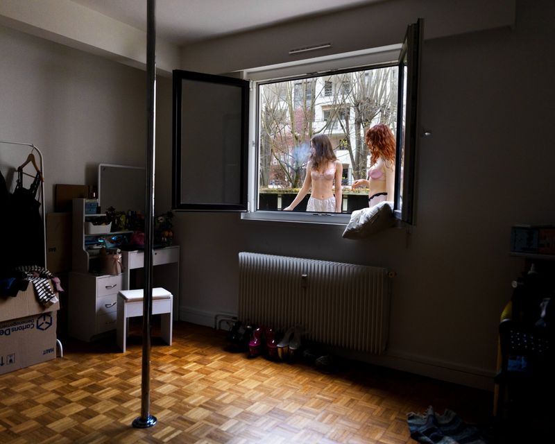 Indoor shot captures two women looking out a window in lingerie, a dance pole stands in the room with visible bed and interior decor.