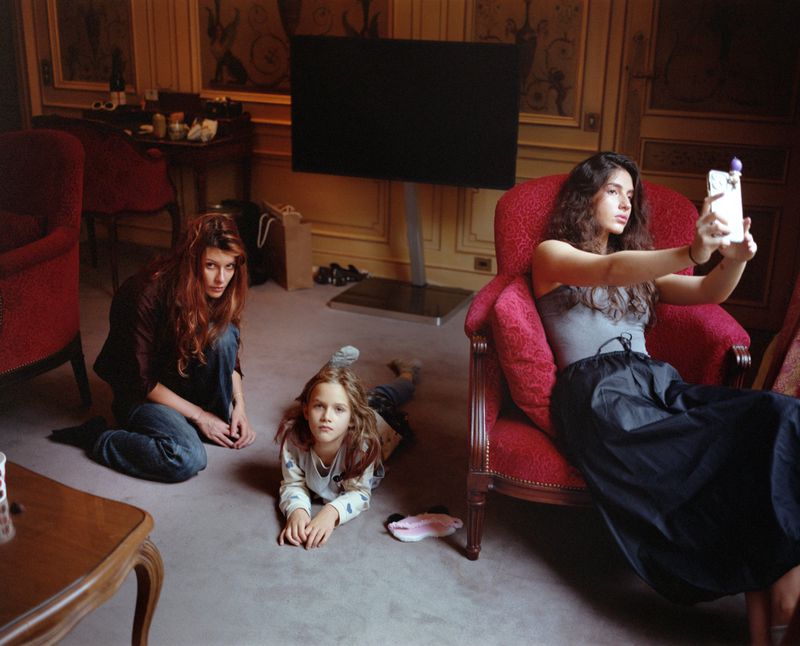 A group of three people are in a room: a young adult taking a selfie, an adult sitting on the floor, and a child lying on the floor.