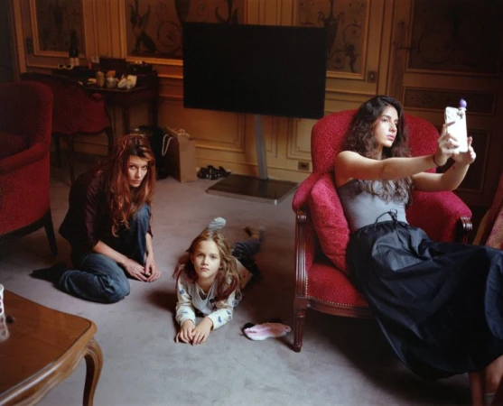 A group of three people are in a room: a young adult taking a selfie, an adult sitting on the floor, and a child lying on the floor.