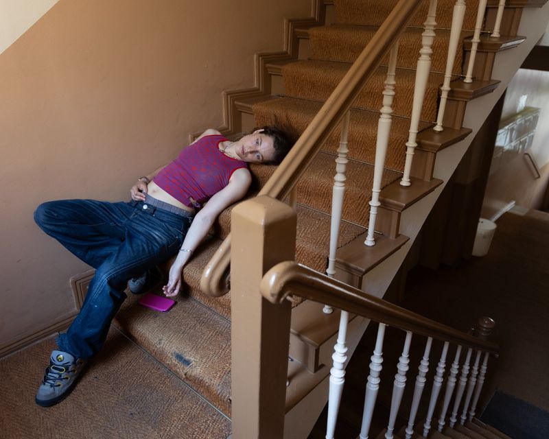 A young adult lies on a carpeted staircase indoors with a cell phone, wearing jeans, a maroon top, and grey sneakers; the stairwell is a light brown.