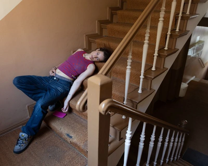 A young adult lies on a carpeted staircase indoors with a cell phone, wearing jeans, a maroon top, and grey sneakers; the stairwell is a light brown.