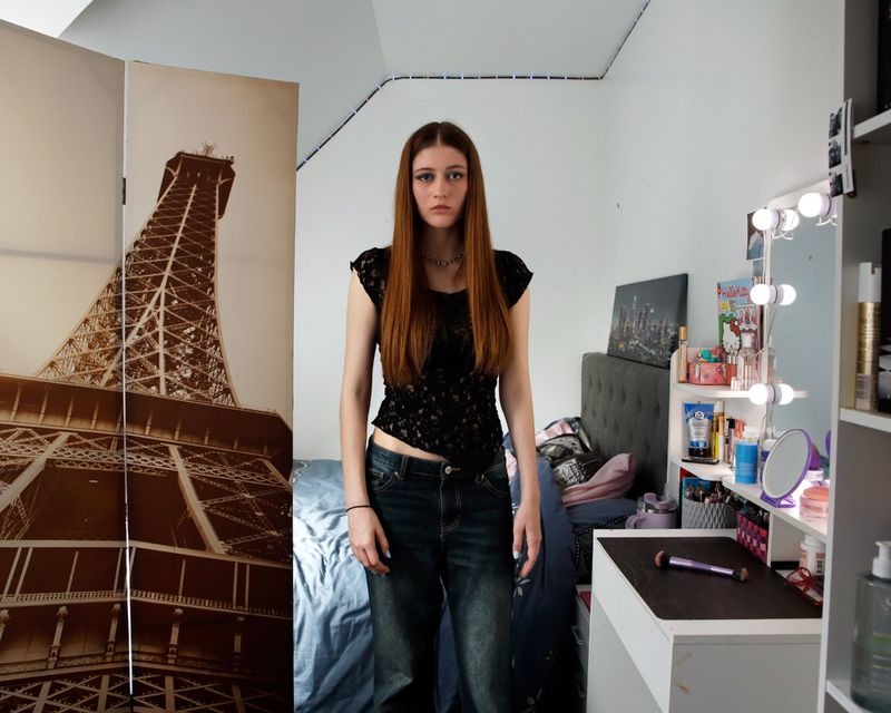 Young woman stands in her bedroom with a screen printed with the Eiffel Tower visible on the left side of the frame.