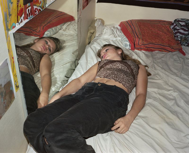 A teenage girl lies on a white bed with patterned pillows, next to a mirror reflecting her resting pose in an indoor setting.