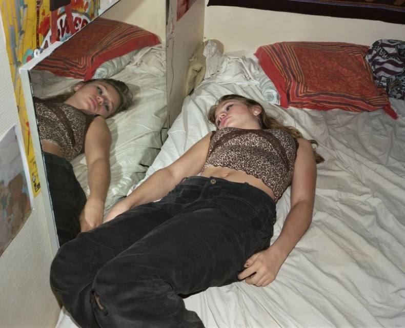 A teenage girl lies on a white bed with patterned pillows, next to a mirror reflecting her resting pose in an indoor setting.