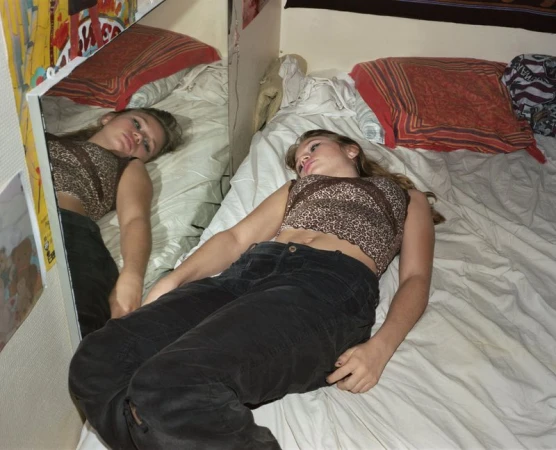 A teenage girl lies on a white bed with patterned pillows, next to a mirror reflecting her resting pose in an indoor setting.