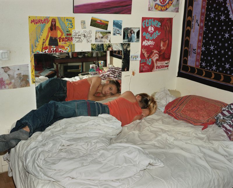 A woman in an orange shirt and jeans lies on a bed with white sheets, in a bedroom with posters on the wall, reflected in a mirror.