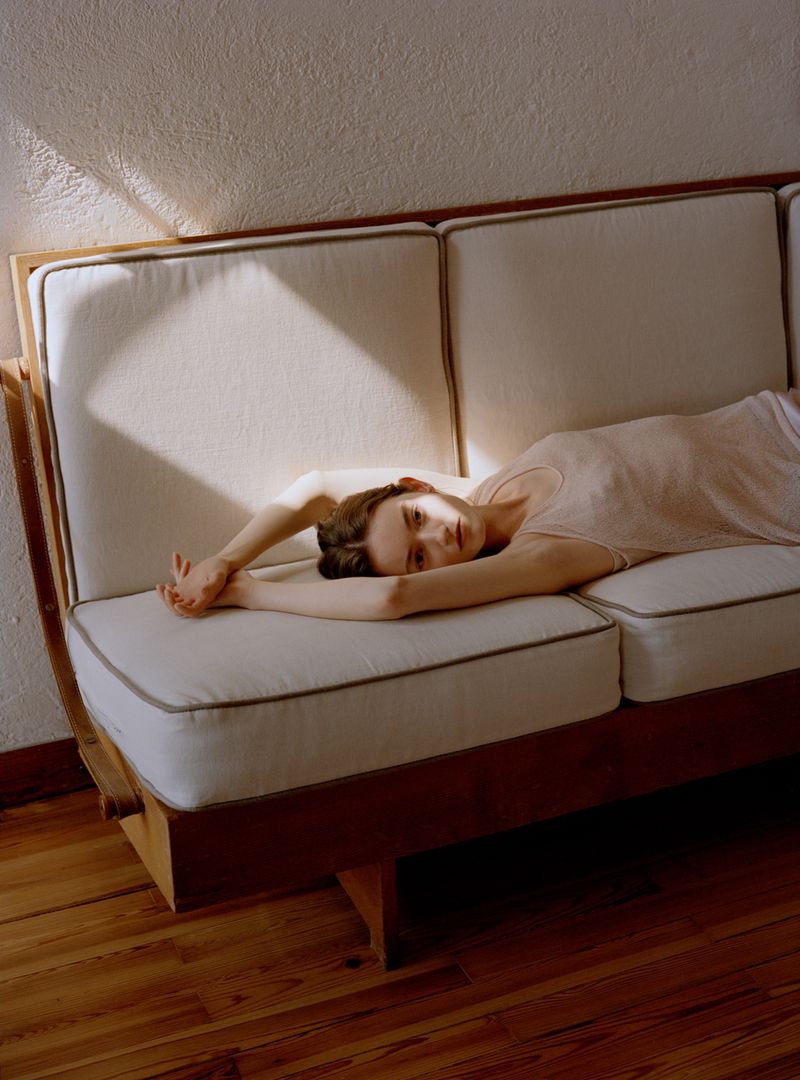 Woman in a beige knit top reclining on a mid-century wooden sofa with cream cushions in natural sunlight.