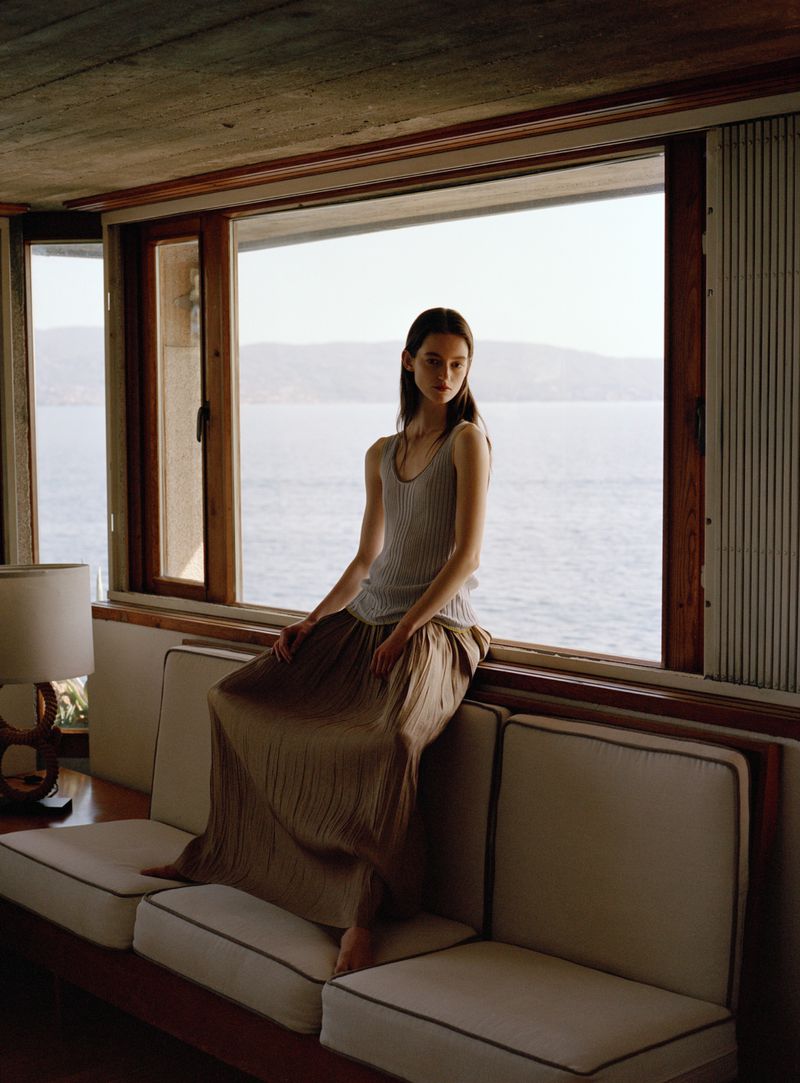 Model seated on a sofa backed against a large window overlooking the sea for Roberto Collina Isole SS26 campaign