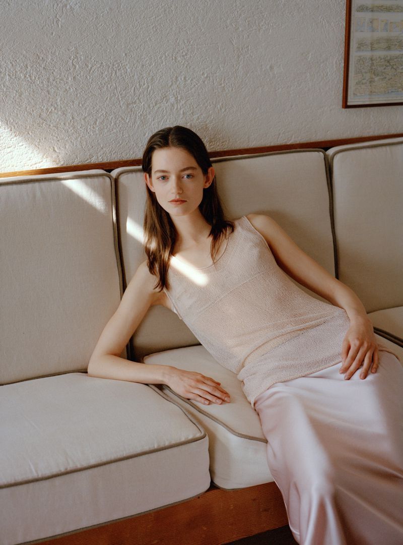 Woman in a pale sheer knit sleeveless top and satin skirt reclining on a cream sofa under natural sunlight.