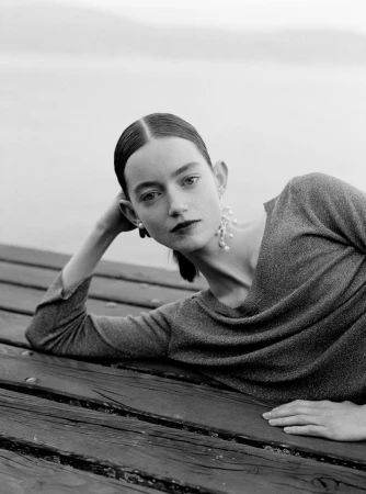 Black and white portrait of a woman in a knit top and pearl earrings lying on a wooden dock by the water.