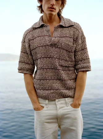 Man wearing a textured knit polo shirt and white trousers standing against an ocean backdrop