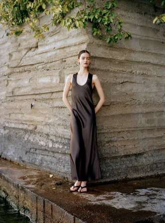 Model standing outdoors wearing a long dark brown slip dress against a textured concrete wall with foliage overhead.