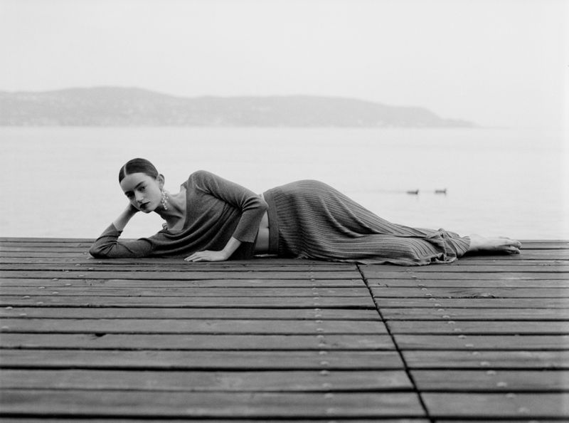 Model lying reclined on a wooden pier wearing a long-sleeve knit top and matching pleated skirt beside a calm lake.