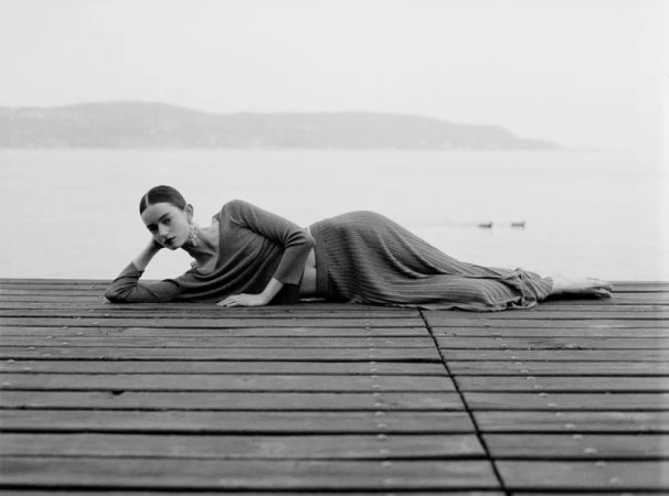 Model lying reclined on a wooden pier wearing a long-sleeve knit top and matching pleated skirt beside a calm lake.