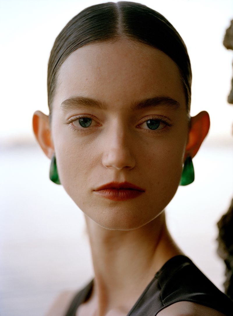 Close-up portrait of a woman with center-parted dark hair wearing green earrings and a black top against a blurred outdoor background