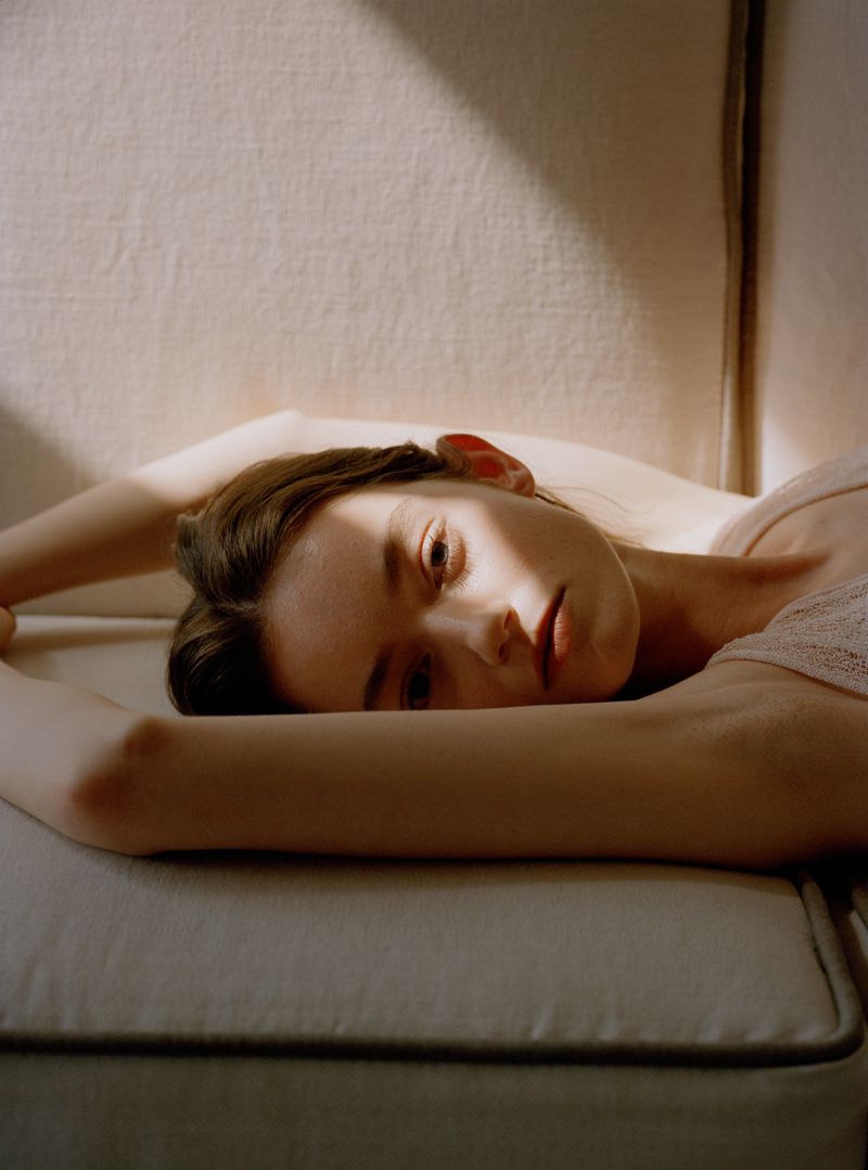 A woman lying on a neutral fabric sofa with direct sunlight casting a diagonal shadow across her face.
