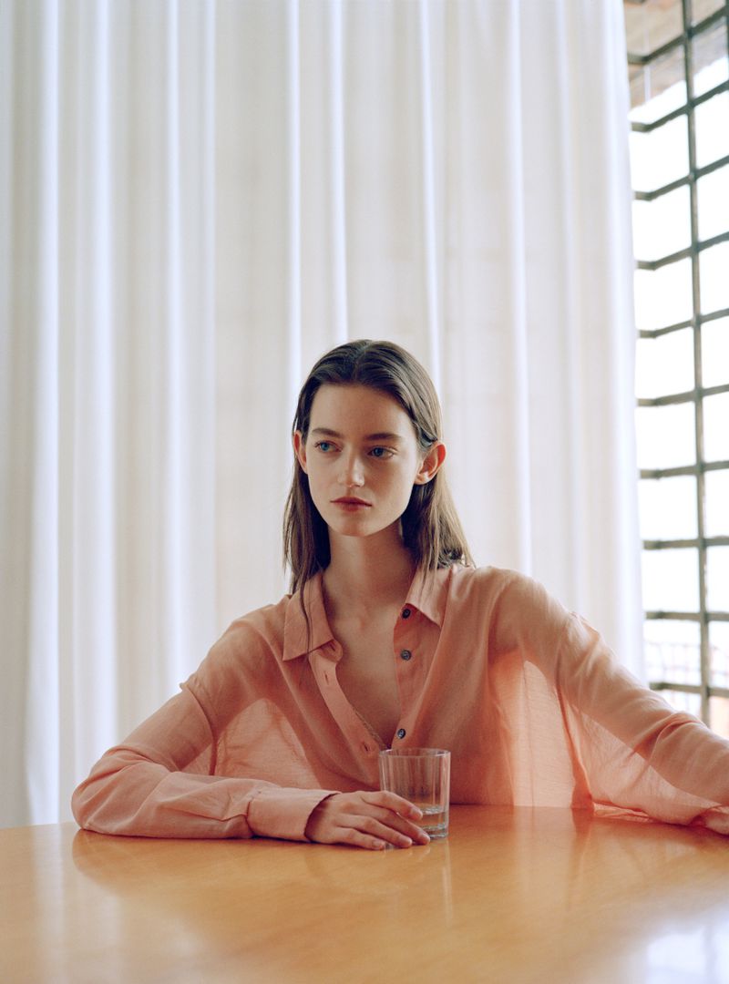 Woman wearing a peach buttoned shirt seated at a wooden table with a glass against white curtains