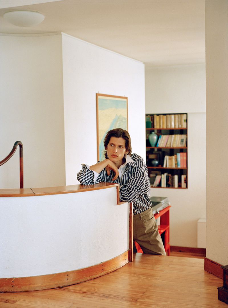 Model resting his chin on his hand while leaning on a curved white interior partition in a striped shirt and khaki pants.