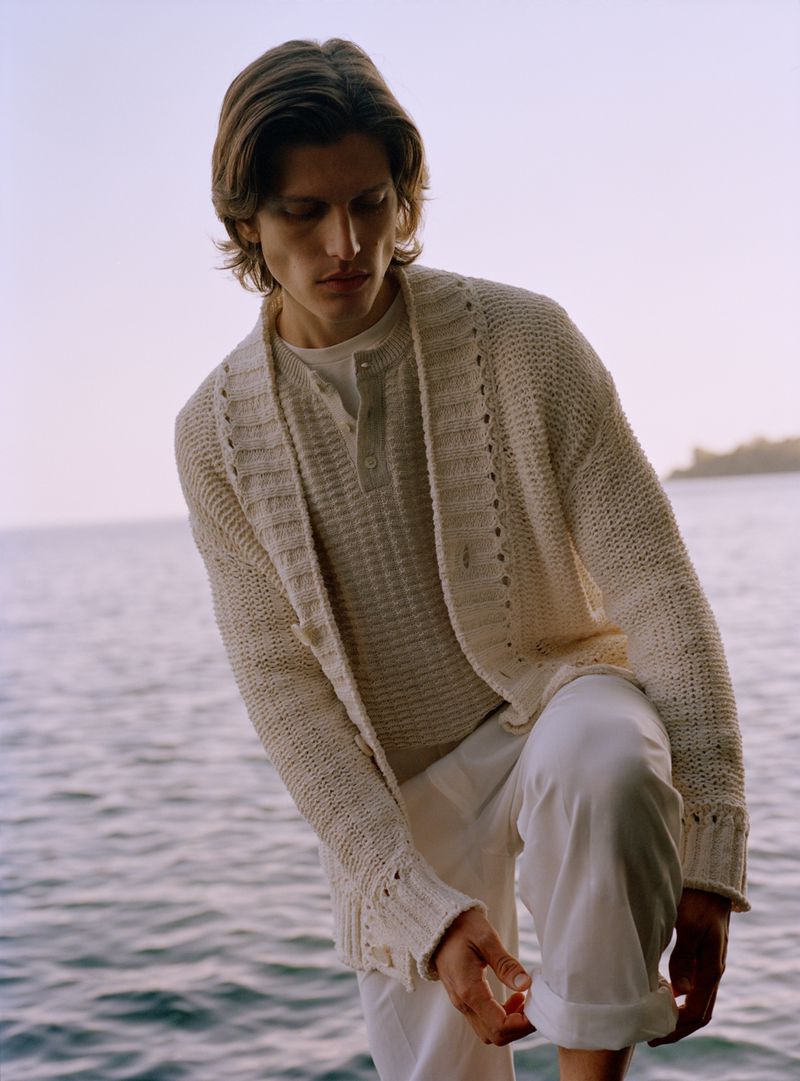 A man in a textured cream knit cardigan and white trousers adjusts his pant cuff against a coastal sea background.