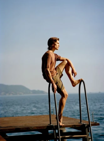 A man in a brown knit shirt and khaki shorts poses barefoot on a wooden dock ladder overlooking the sea.
