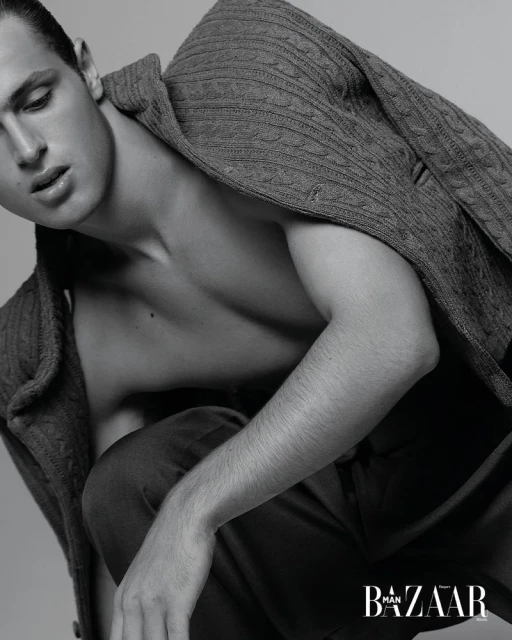 Portrait of a shirtless man in a gray sweater and trousers, posing in front of a neutral backdrop, with Harper's Bazaar magazine logo visible.