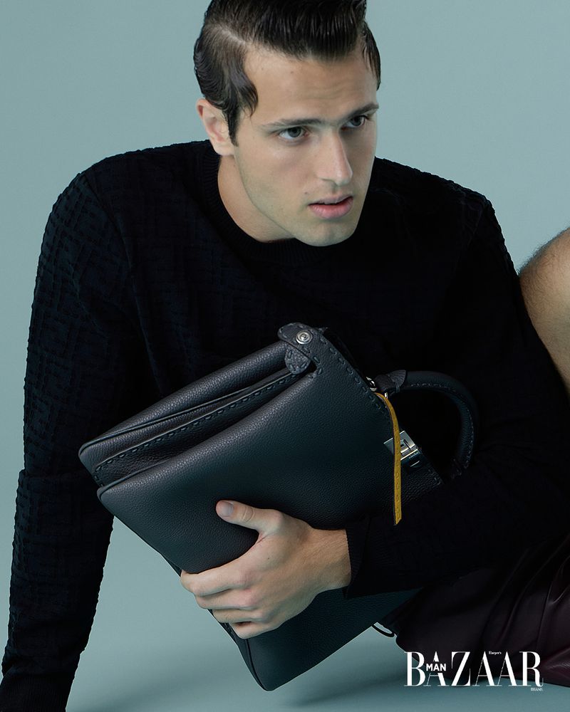 Masculine fashion portrait: dark-haired man in a black sweater holds a leather bag with 'Bazaar Man' logo visible in the bottom corner.