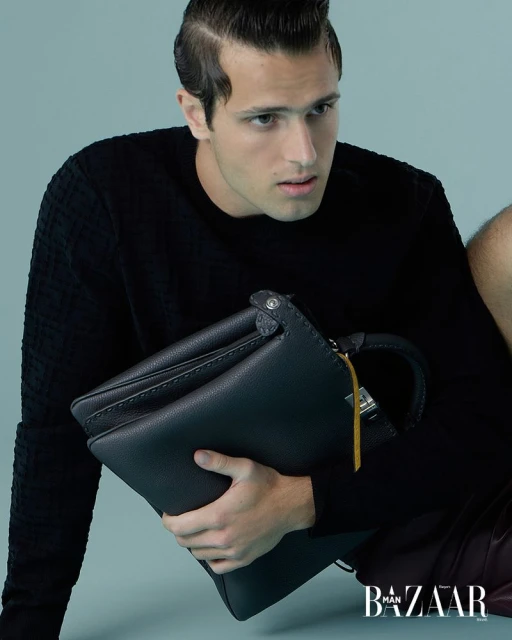 Masculine fashion portrait: dark-haired man in a black sweater holds a leather bag with 'Bazaar Man' logo visible in the bottom corner.