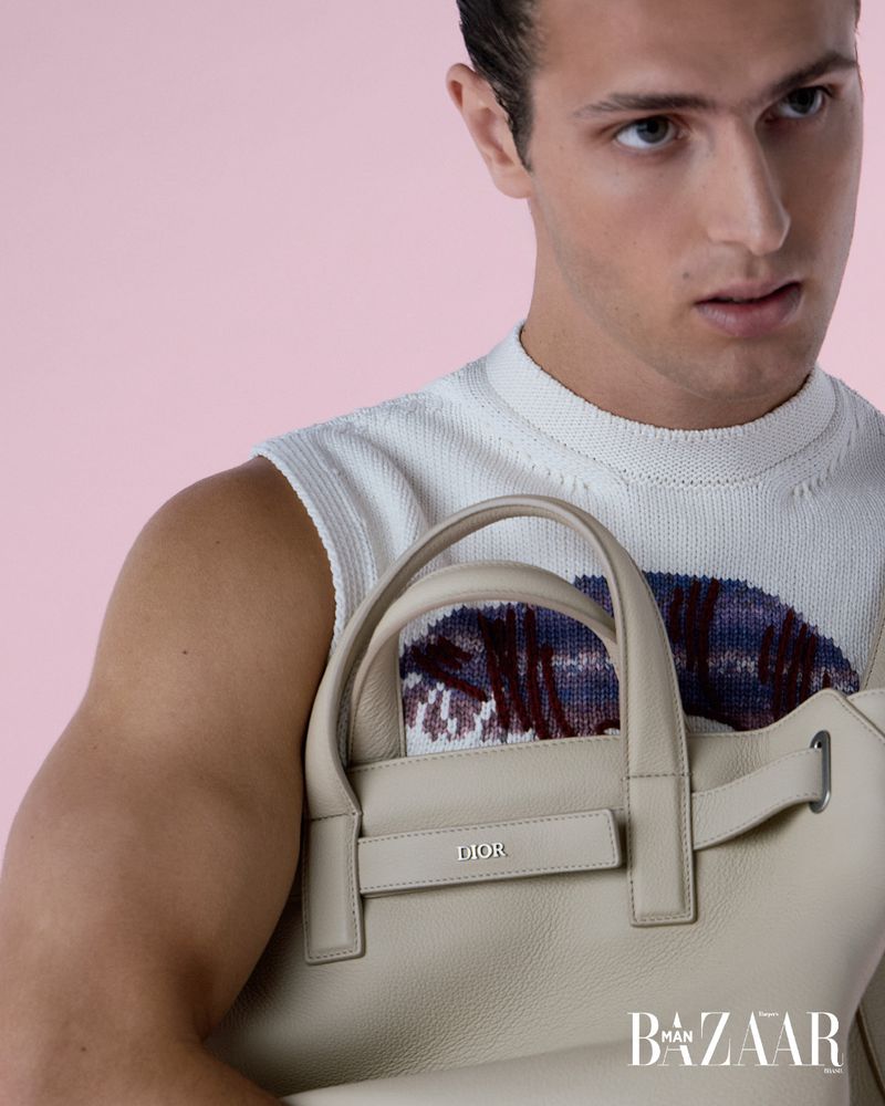 Man poses with Dior handbag against a pink backdrop, wearing a white patterned top in a studio shot for a Man Bazaar ad.