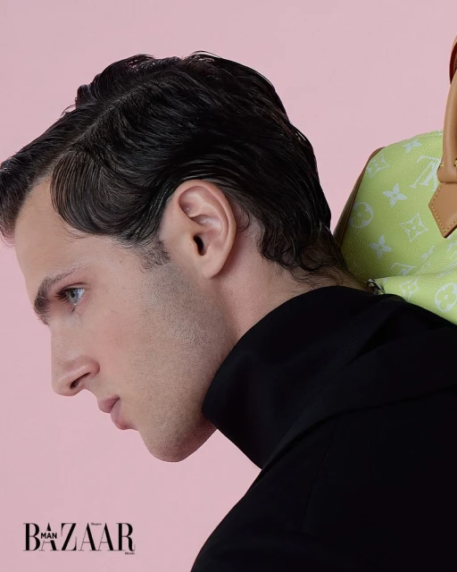 Close-up profile portrait of a dark-haired man wearing a black turtleneck sweater and a lime green bag on a pink background.