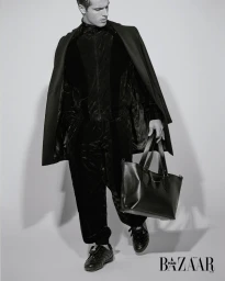 Monochrome fashion editorial image of a male model wearing black velvet attire and holding a leather tote bag against a white backdrop.