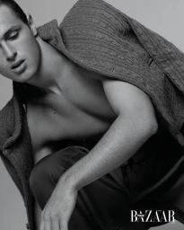 Model Teo Toranzo crouching with a cable-knit sweater draped over his back in a black and white fashion editorial studio portrait.