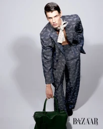 Model Teo Toranzo in a printed suit holding a dark green leather bag against a studio backdrop for Harper's Bazaar Brazil.