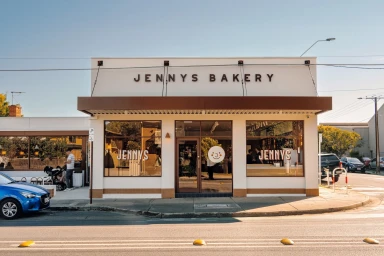 Exterior storefront of Jenny's Bakery featuring large display windows, brown awnings, and clean white facade along a paved street.