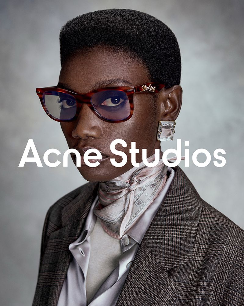 Model Fatou Kebbeh in tortoiseshell glasses, silk neck scarf, and brown plaid blazer against a grey textured studio backdrop.