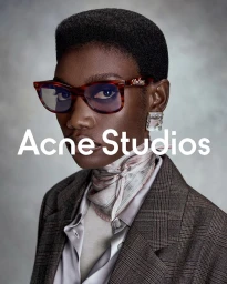 Model Fatou Kebbeh in tortoiseshell glasses, silk neck scarf, and brown plaid blazer against a grey textured studio backdrop.