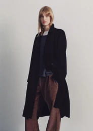 Model wearing a long black tailored coat over wide-leg brown trousers against a pale grey seamless studio backdrop.