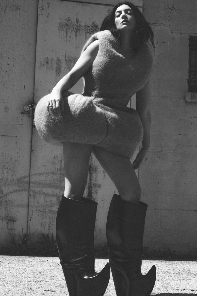 Monochrome fashion portrait of a woman in a unique furry dress and black boots posing against a weathered metal wall outdoors.