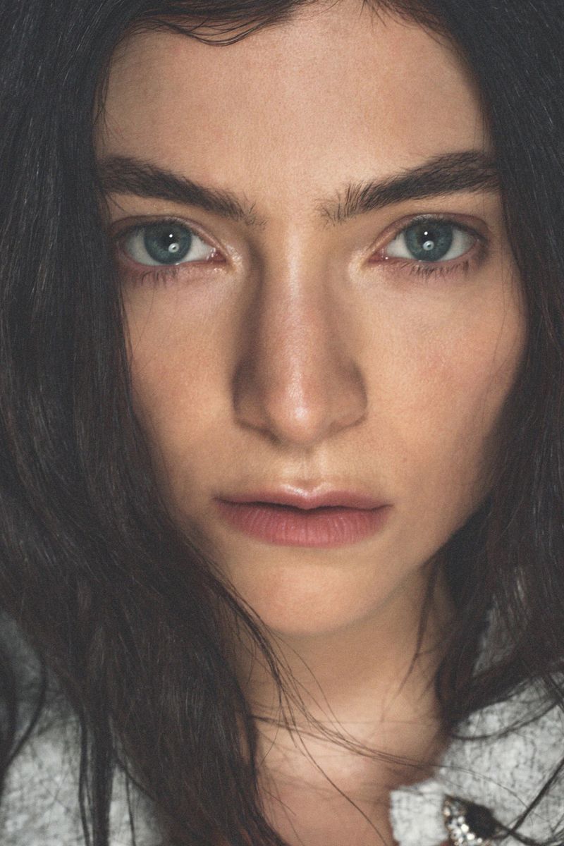 Close-up portrait of a young adult woman with blue eyes and dark brown hair, wearing a grey jacket, looking directly at the camera.