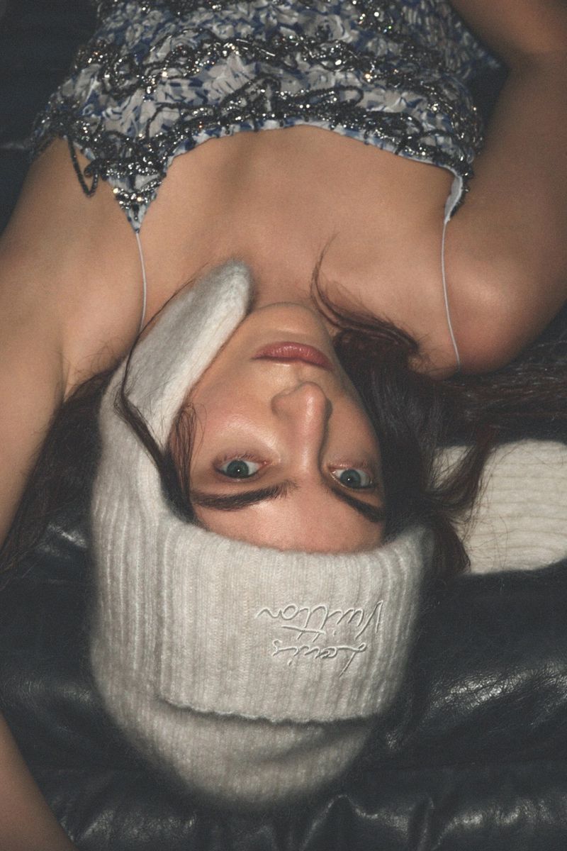 Close-up of an adult female model with long brown hair, wearing a sequined top and a knitted rabbit-ear hat, lying on a dark surface.