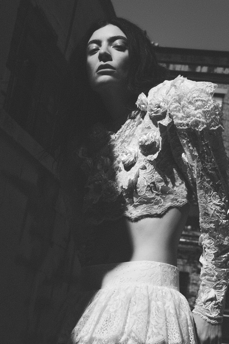 Black and white portrait of Lorde wearing an ornate white floral lace cropped top and matching skirt outdoors in directional sunlight.