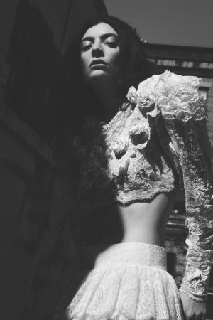 Black and white portrait of Lorde wearing an ornate white floral lace cropped top and matching skirt outdoors in directional sunlight.