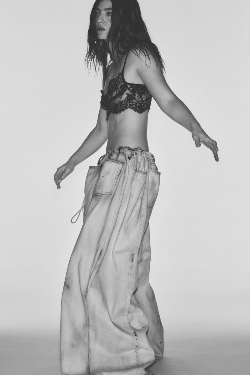 A stylish young adult female model poses in a photo studio wearing a black lace bra and baggy jeans in a black and white medium shot.
