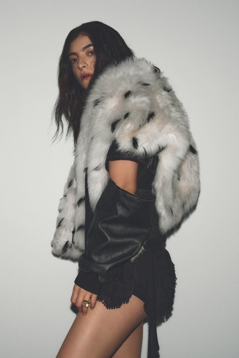 Elegant woman in a leather jacket and fur stole poses in front of a neutral background, showcasing fashion and personal style.