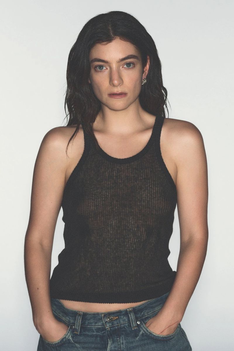 Close-up studio shot of a young adult female posing with hands in jean pockets against a white backdrop, wearing a black ribbed tank top.