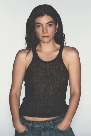 Close-up studio shot of a young adult female posing with hands in jean pockets against a white backdrop, wearing a black ribbed tank top.